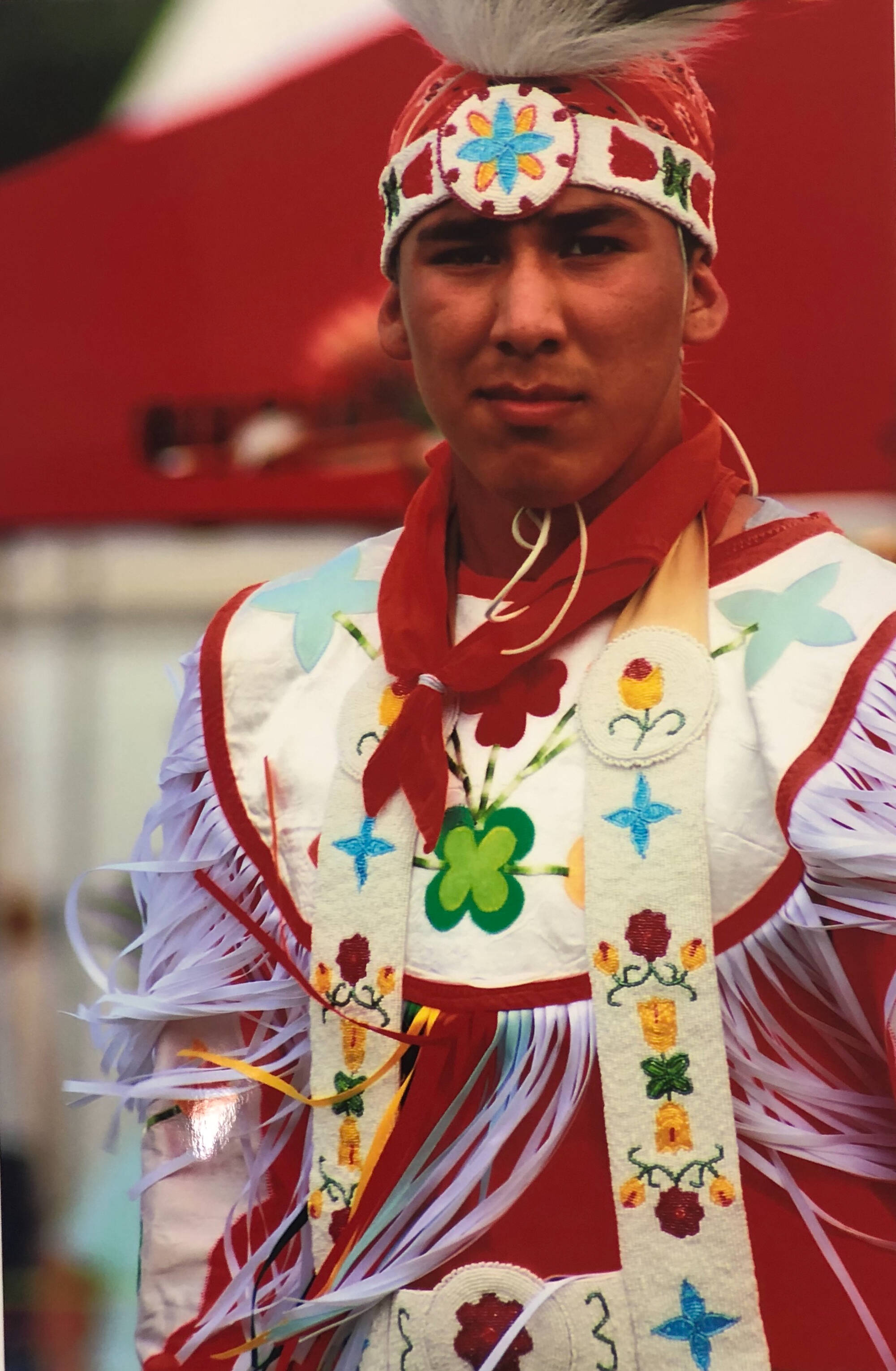 Colored photograph of a Native American man dressed in traditional regalia. His outfit is mostly white and red.